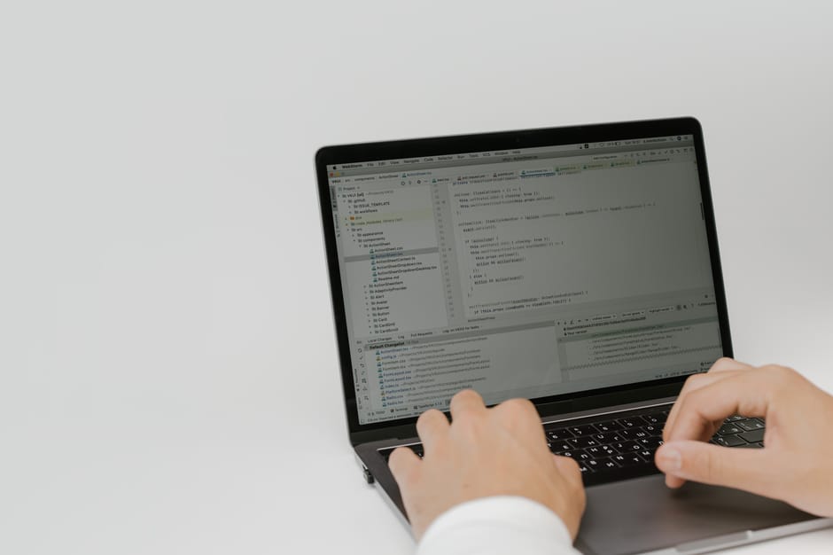 A person working on a laptop with a software development environment open on the screen. The screen displays multiple code files, with scroll bars indicating ongoing editing or review. The person's ha